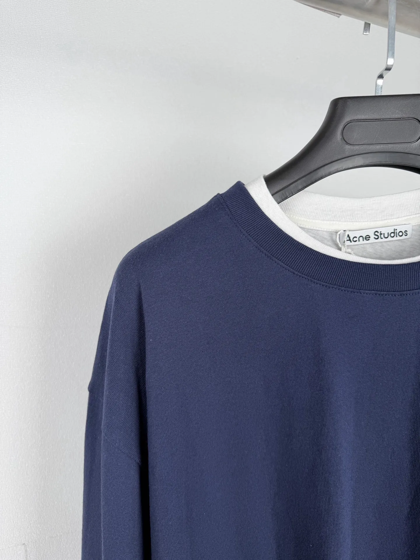ACNE Studios Logo Layered Sweatshirt The outer body is made of 12-count roving to give the cloth a clear twill texture and a dry and stiff skeleton. The surface has a slight natural bamboo texture, which is wrinkle-resistant and stylish. It can firmly support the loose silhouette of the sweatshirt without collapse. The bottom surface of the lining is introduced with 32-count snowflake cotton, and a small amount of dark or different-colored cotton fibers are mixed into the yarn. After weaving, the cloth surface presents a fine and uniform granular pitted texture, which enriches the tactile layer and enhances the breathable moisture-repelling property. The two fabrics are combined through a fake two-piece structure, which not only retains the rough and crisp visual effect of the outer layer, but also keeps the skin-adjacent surface delicate, soft and light, avoiding the sultry constraints caused by a single heavy fabric. The embroidered logo on the chest uses a high-density flat embroidery stitch method, using coarse cotton as the base. The embroidery thread is tightly covered, and the edges are trimmed with stitches to ensure that the logo is clear and three-dimensional on the rough-textured cloth without revealing the background color. The stitches outline the outline with short overlapping stitches. Overlapping stitches are the characteristic craft technique. The embroidery needles are staggered back and forth as they move, forming a fine and rhythmic line texture.